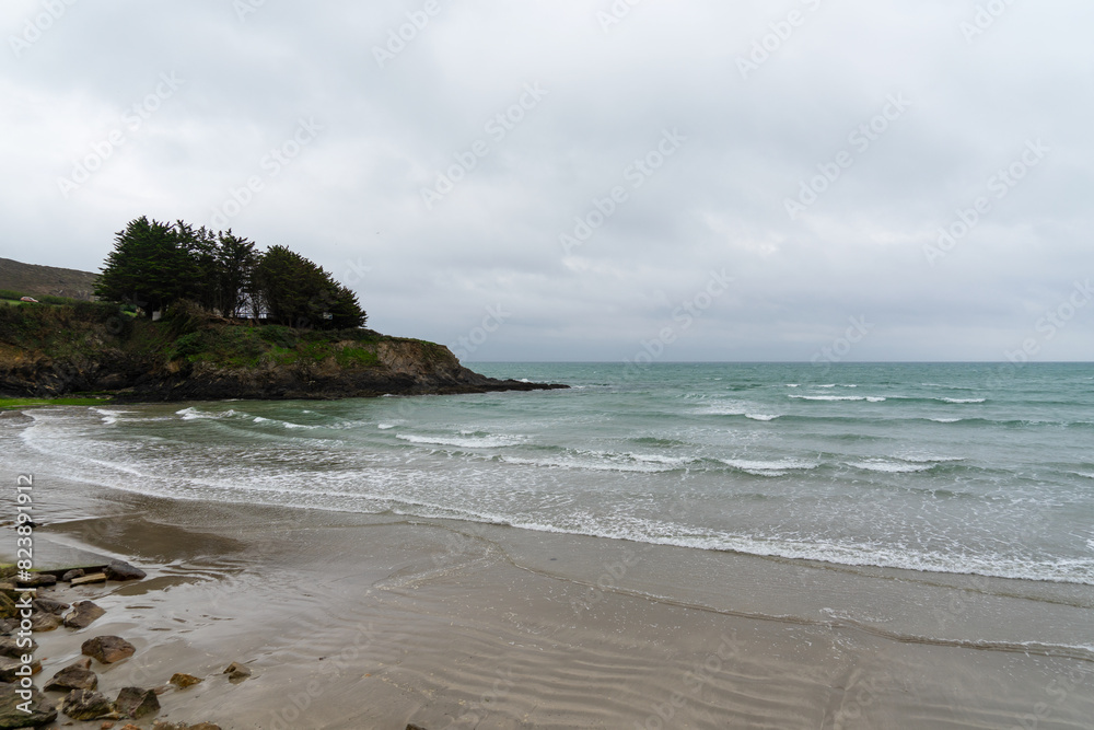 Plage de la presqu'île de Crozon, où les eaux émeraudes de la mer d'Iroise rencontrent le sable, sous un ciel couvert où se dessinent les nuances grises des nuages, l'écume blanche ourle le rivage.