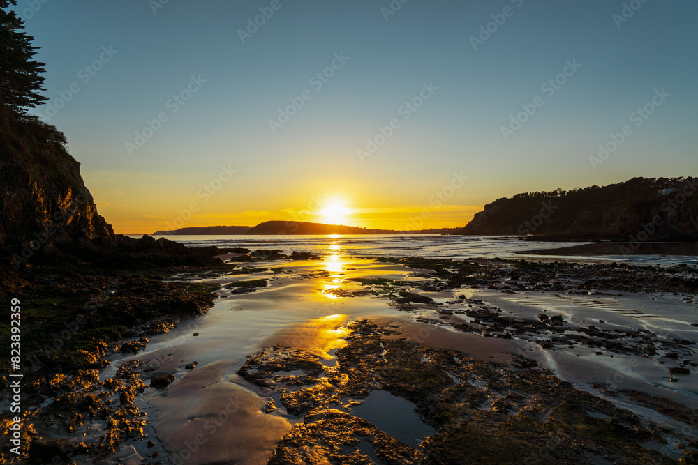 Obraz premium Le soleil se couche sur une plage de la presqu'île de Crozon, en Bretagne, teintant le ciel de jaune et baignant le sable doré d'une lumière douce, tandis que l'océan murmure doucement.