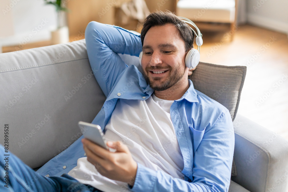 A man is sitting on a couch while listening to music on his phone. He appears relaxed and engaged in the music playing through his headphones. A man is sitting on a couch while listening to music on his phone. He appears relaxed and engaged in the music playing through his headphones.