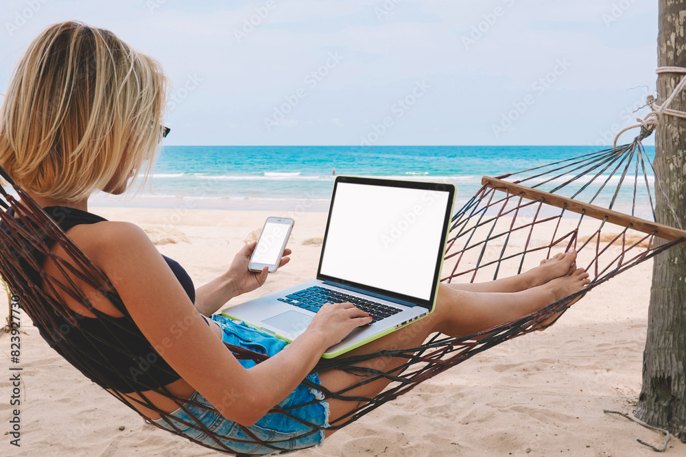 Female freelancer entrepreneur lying on hammock and doing remote business work at modern laptop computer with blank screen area for web page on smartphone during summer vacation on tropical seashore