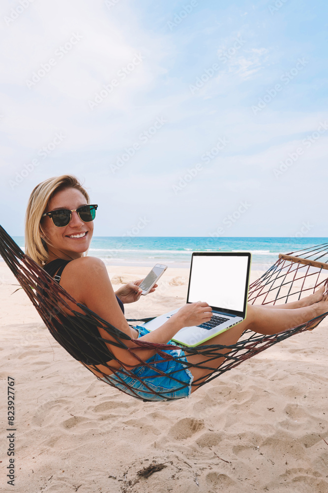 Happy successful female work freelance at laptop with blank screen area for web content while lying on hammock and typing sms message in roaming on smartphone during summer vacations on tropic island
