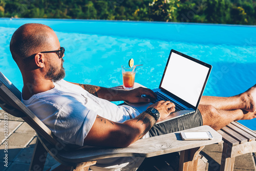 Businessman enjoying hot tour during summer resort lying on sunbed with cocktail near pool while earning money doing distance job at netbook with blank copy space screen for your advertising content