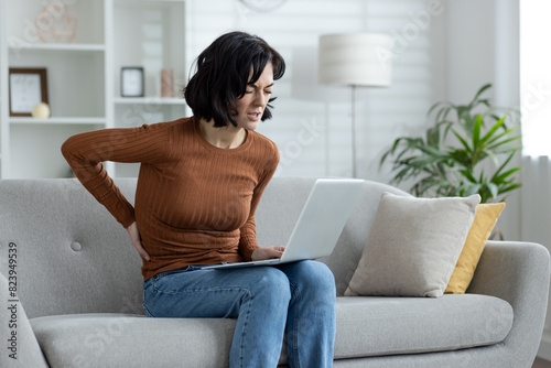 Young woman suffering from back pain, sitting at home on the sofa, working and studying remotely via laptop, holding her hand on her lower back