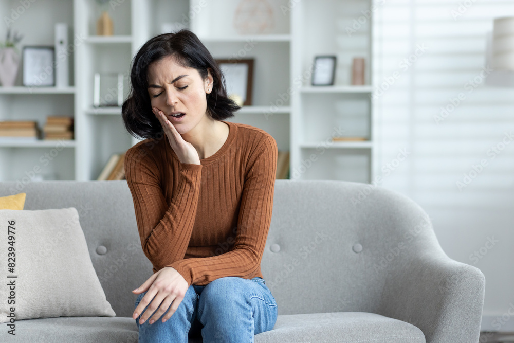 A sick young woman sits at home on the sofa and holds her hand to her cheek, grimaced from severe toothache, needs medical help from a dentist A sick young woman sits at home on the sofa and holds her hand to her cheek, grimaced from severe toothache, needs medical help from a dentist