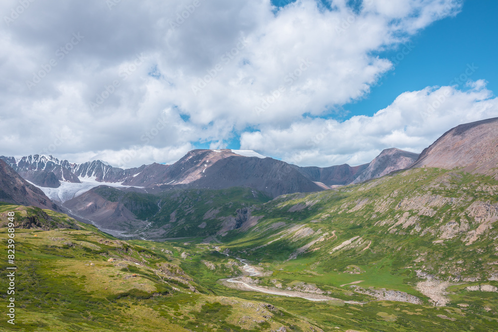 Wide green alpine valley with snake river among hills and rocks with vast view to large snow mountain top and big glacier tongue under cloudy sky. Serpentine river in high mountains. Shadows of clouds