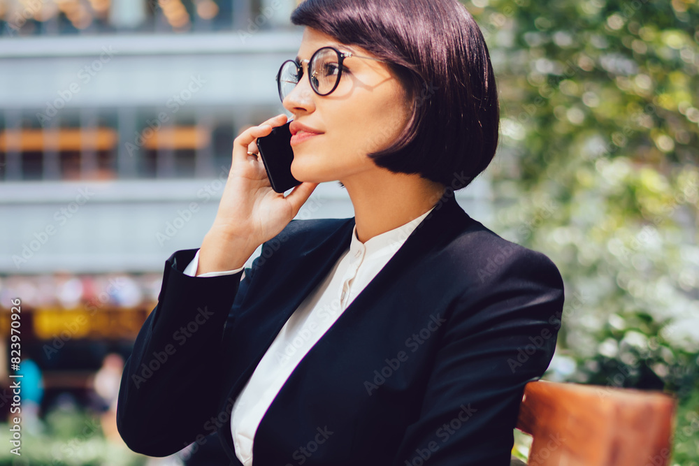 Female financial director of company in eyeglasses having phone conversation about work sitting on bench with coffee to go near office building.Manager in elegant wear talking on mobile during break