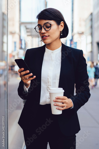 Confident female lawyer in formal wear strolling in metropolis and transferring money on phone.Serious owner holding takeaway coffee cup walking on business district and messaging on smartphone
