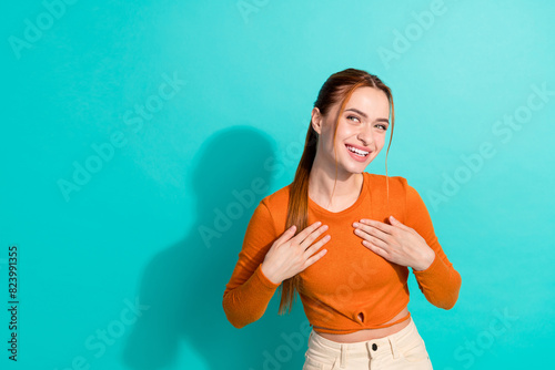 Photo of positive cunning tricky girl with ginger hairdo dressed orange shirt look at offer empty space isolated on teal color background photo