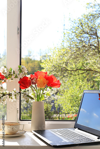 A cup of tea, a computer, tulip flowers in a vase on a sunny window. The concept of home coziness, comfort and home office. Computer mobility and healthy lifestyle, selective focus..