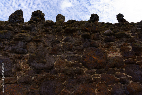 Old ruined battlement or crenellated wall, rough-textured wall constructed from irregularly shaped stones in shades of brown and black, ancient traditional Building technique, low-angle shot