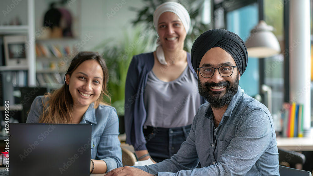 A smiling Indian man wearing a turban poses with two female colleagues in front of a laptop. A smiling Indian man wearing a turban poses with two female colleagues in front of a laptop.
