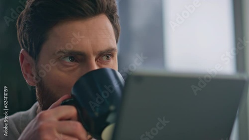 Closeup guy drinking coffee reading tablet at morning apartment. Man holding tab