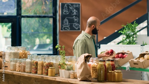 Client entering local grocery store to buy organic fruits and vegetables, looking at pasta and grains stored in sustainable nonpolluting glass jars. Middle eastern man advertising healthy eating. photo