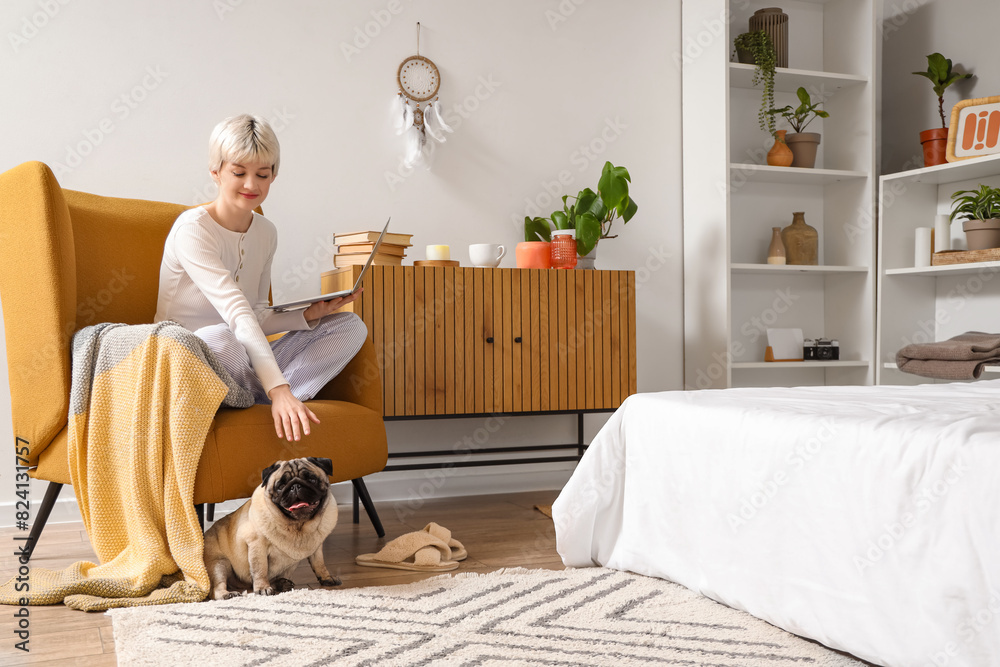 Young woman with pug dog using laptop in armchair at home Young woman with pug dog using laptop in armchair at home