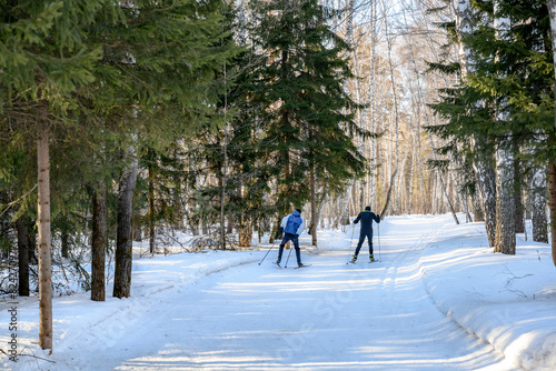 Two skiers are running on a ski track in a winter forest with snowdrifts under the bright sun