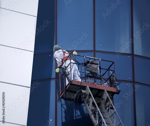 An installer dressed in a uniform and a safety harness cleans the glass facades while standing on a construction winch