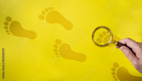Human hand holding a magnifying glass looking at human footprints on a yellow background. Concept of searching for traces of theft or tracking.