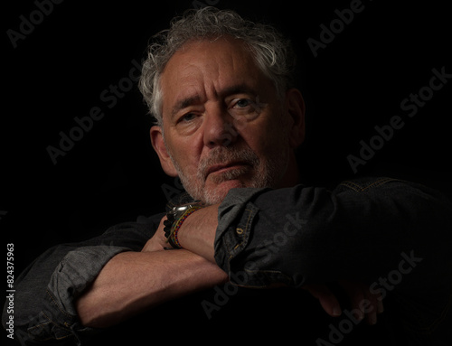 Low key color portrait of an attractive older Caucasian man with grey hair and beard. Looking at camera with serious or pensive expression, leaning on his arms.  