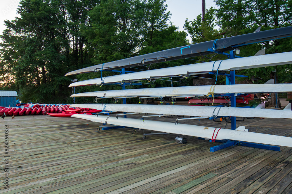 A row of rowing boats is neatly arranged on racks along a wooden boat dock, while kayaks are meticulously organized on the ground below. The wooden deck.