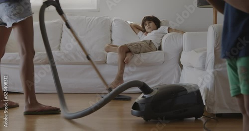 A mother vacuums the living room while her young son watches from the couch, appearing bored and uninterested.