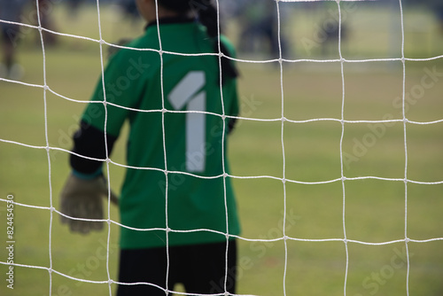 Close up football goal net, seeing a young girl soccer goalkeeper watching her teammates playing. Youth Soccer game on a sunny summer school tournament Day. A football match going on in a background.