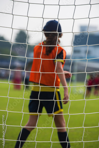 Close up football goal net, seeing a young girl soccer goalkeeper watching her teammates playing. Youth Soccer game on a sunny summer school tournament Day. A football match going on in a background.