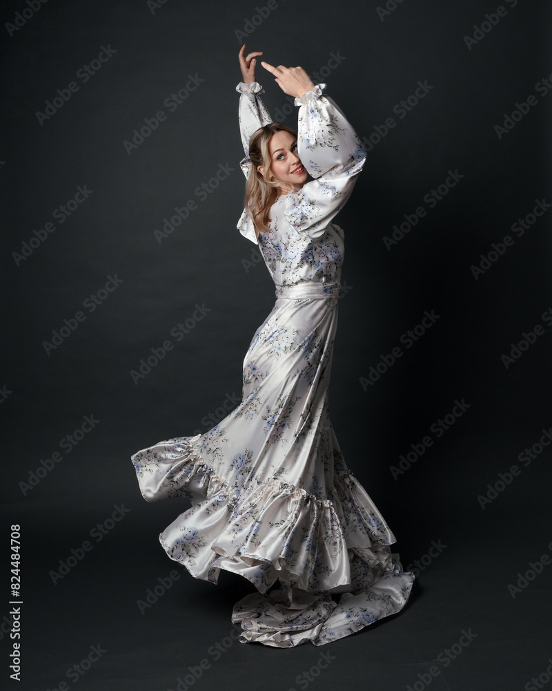 portrait of beautiful blonde female model wearing romantic historical gown white bridal floral pattern. standing pose running away from camera with flowing silk skirt. isolated dark studio background.