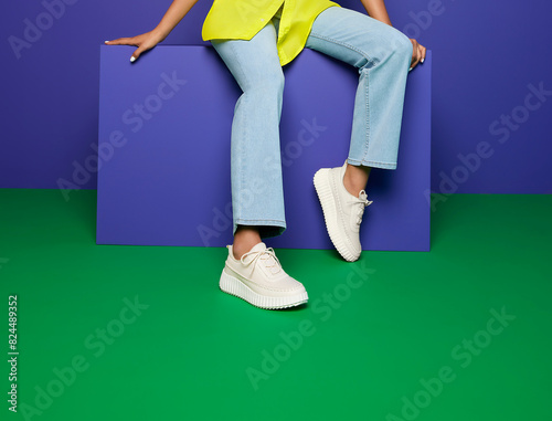 Young woman modeling trendy cream-colored lace-up sneakers with a chunky tread sole, seated on a purple block, a green and purple background. dressed in light blue jeans, yellow top, casual footwear.