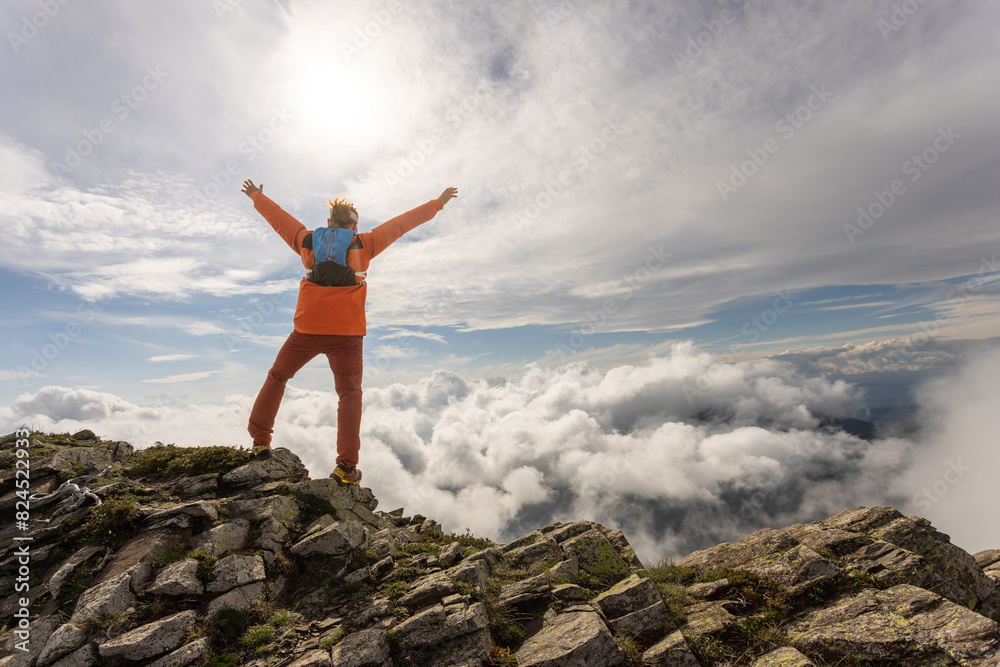 A man in an orange jacket is standing on a mountain top, looking up at the sky A man in an orange jacket is standing on a mountain top, looking up at the sky