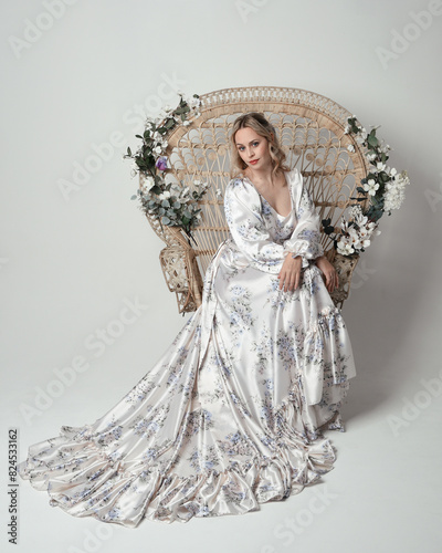 portrait of beautiful blonde female model wearing romantic historical white bridal gown. sitting pose on floral Peacock Throne chair with flowing silk skirt. isolated dark studio background.