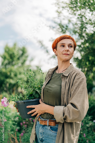 Beautiful adult redhead woman in her garden holding flower pot with petunia wearing casual style and short haircut wellness and nature calm