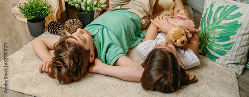 Banner of serene boy and little girl embracing stuffed dog resting surrounded by cushions and plants at home. Two children relaxing lying over rug in a warm and playful ambiance at home.