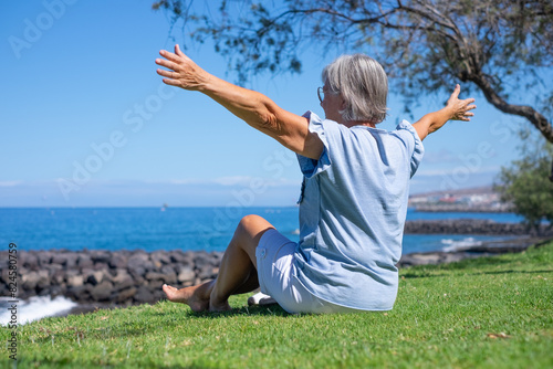 Back view of senior woman sitting barefoot in meadow fae the sea looking at the horizon with open arms, relaxed senior lady enjoying free time summer vacation or retirement #824580759