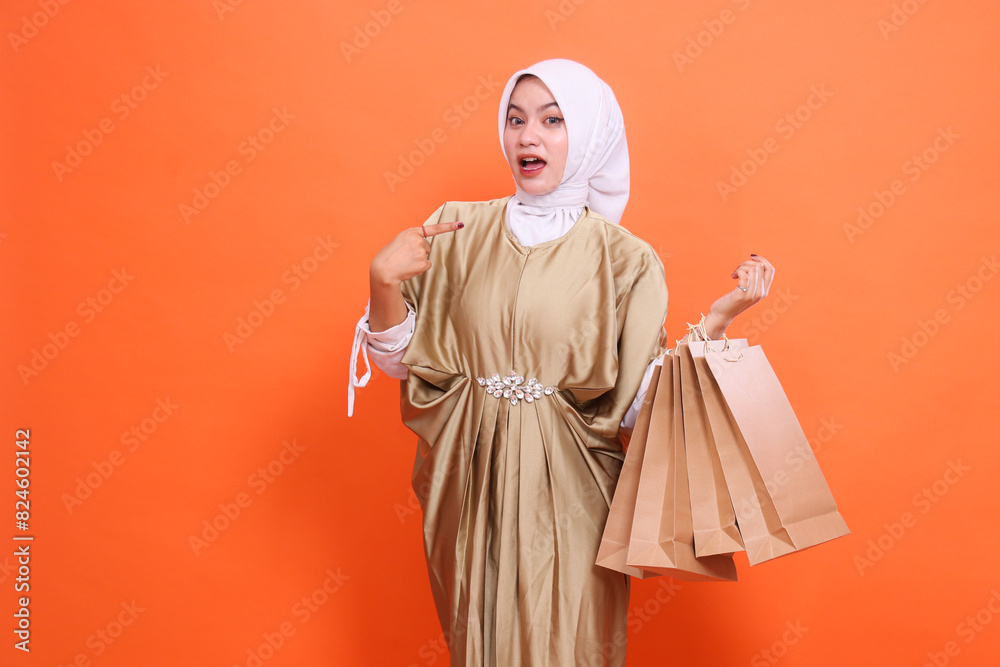 shocked mature Asian woman carrying a brown paper bag containing shopping items while pointing at it wearing a kaftan Muslim dress with a hijab,on an orange background. Fashion, transaction, promotion