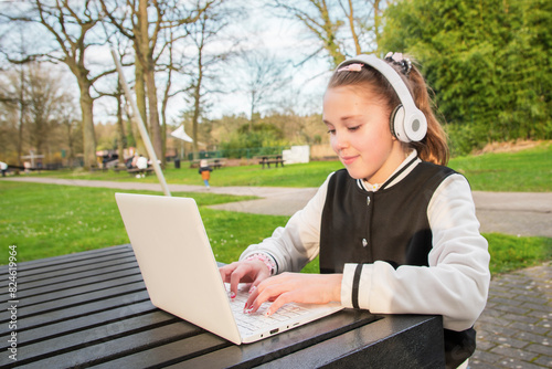 Teenager girl sits at a table with a laptop, white headphones and cup of tee outside photo