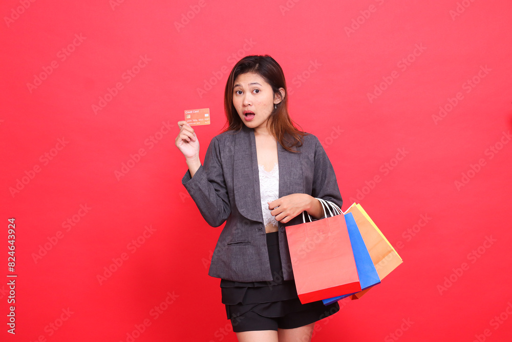 Asian office woman gestures in surprise at the camera carrying a shopping paper bag and holding a credit debit card wearing a formal suit with a red background. for transaction, lifestyle and business Asian office woman gestures in surprise at the camera carrying a shopping paper bag and holding a credit debit card wearing a formal suit with a red background. for transaction, lifestyle and business