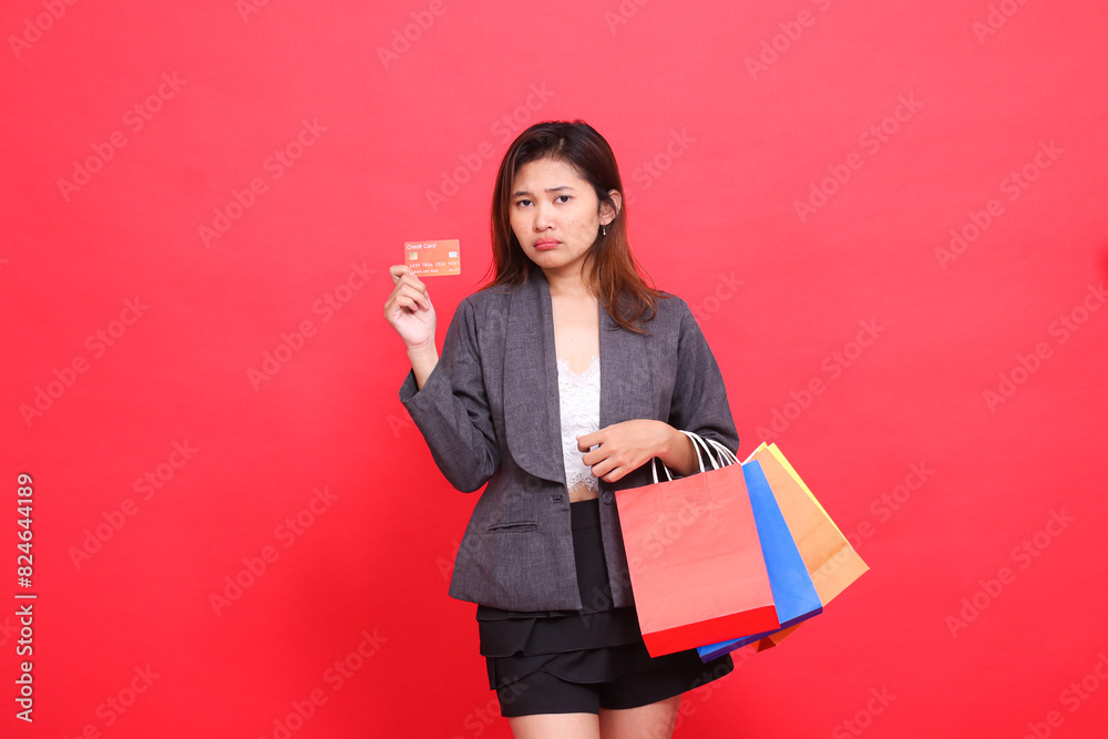 sad Asian office woman gestures to the camera carrying a shopping paper bag and holding a credit debit card wearing a formal suit on a red background. for transaction, lifestyle and business sad Asian office woman gestures to the camera carrying a shopping paper bag and holding a credit debit card wearing a formal suit on a red background. for transaction, lifestyle and business