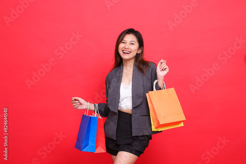 Expression of a happy Asian office woman carrying a shopping paper bag wearing a formal suit on a red background. for commercial, lifestyle and sales concepts photo