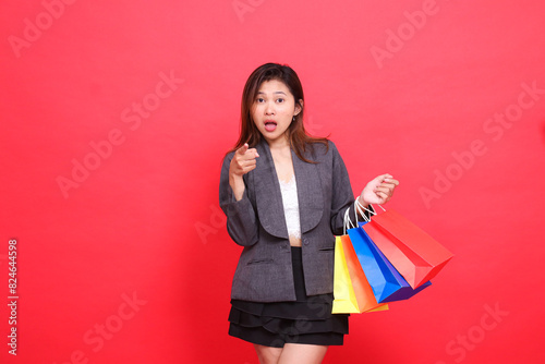 gesture of surprised asian businesswoman carrying shopping paper bag, hand showing you wearing formal jacket on red background. for commercial, lifestyle and sales concepts photo