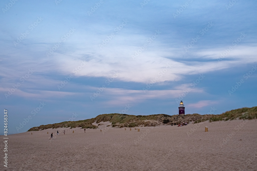 In the soft light of the early evening, several individuals take a leisurely stroll along the expansive sandy beach, with the dunes rising gently in the background. A prominent lighthouse, its vivid
