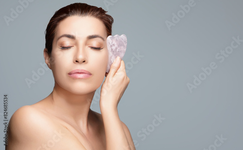 Woman practicing facial massage with gua sha stone on isolated gray background