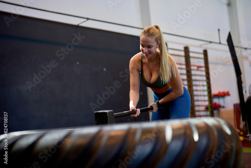 Young caucasian woman hitting a large tire with a sledgehammer in the gym.