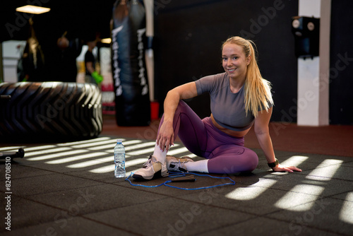 Young sporty woman posing for a photo after completing her workout, sitting on the gym floor with a water bottle and jump rope.