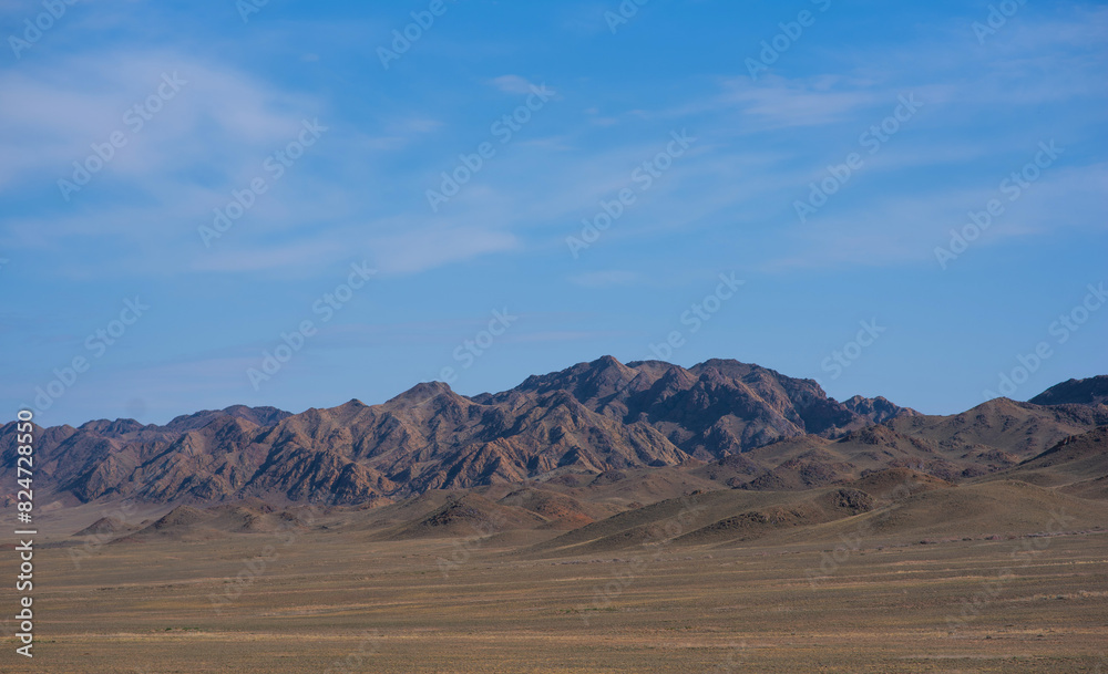 a rugged mountain range under a vast blue sky, with undulating hills in the foreground and sharp, rocky peaks in the distance creating a dramatic landscape