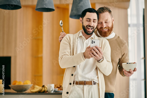 Two men happily smiling while holding a cell phone in a modern apartment setting.