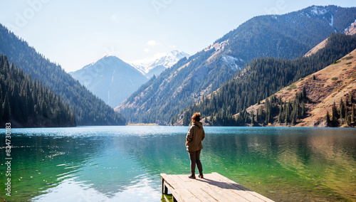 a adult woman standing on a dock overlooking a tranquil alpine lake, gazing at the snow-covered mountain peaks and surrounding dense pine forests under a clear blue sky