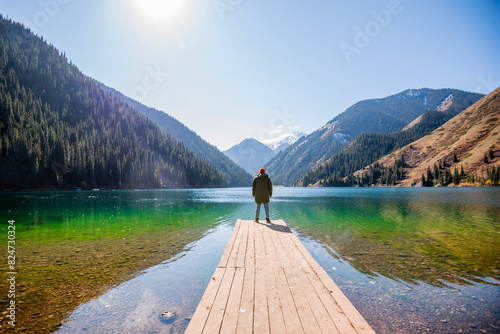 a man standing at the end of a wooden pier, looking out over a clear alpine lake towards towering, snow-capped mountains and dense pine forests under a bright sunlit sky