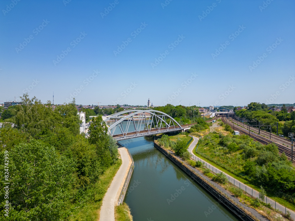 Aerial view of a bridge over a river with greenery and urban surroundings on a clear, sunny day in a scenic photograph