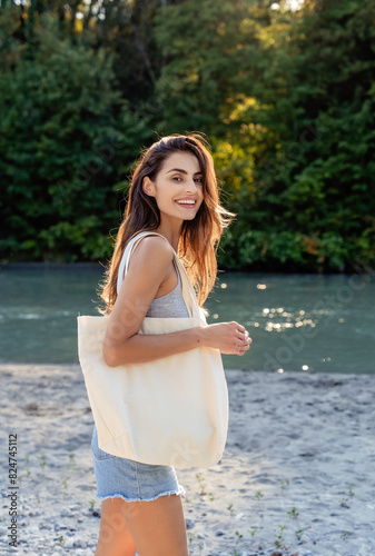 Young happy smiling woman with long hair walking on sandy river beach in countryside with white cotton eco friendly tote bag looking at camera. Mock up. Summer vacation on sunny day.