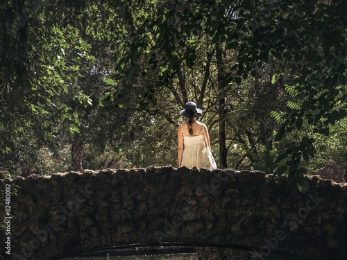 A young woman girl in a dress and hat from behind back standing on a rock stone bridge in a forest park during summer photo